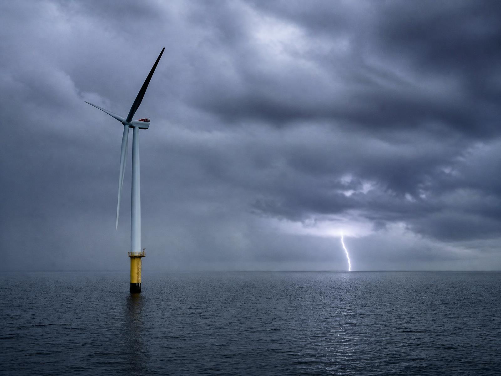 Offshore wind turbine standing safely as lightning strikes the sea in the distance